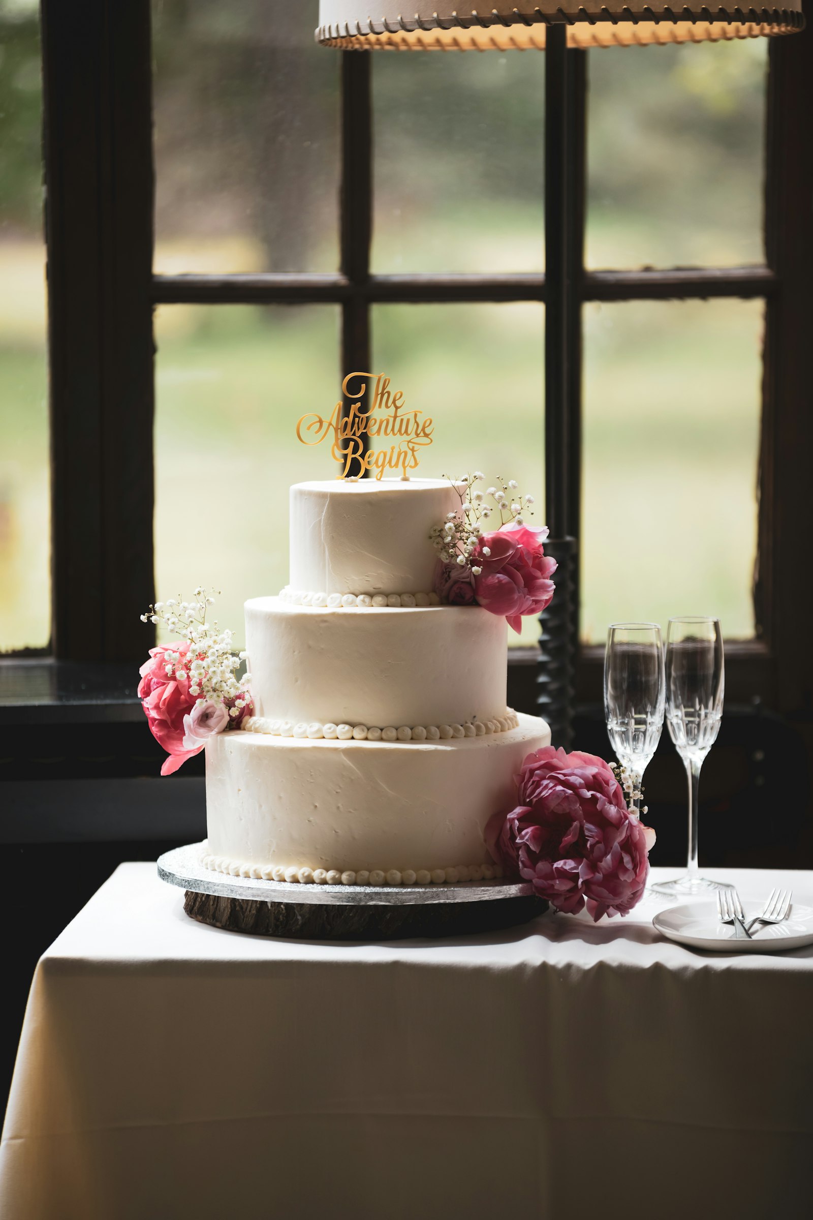 Three-tier wedding cake decorated with fresh pink flowers, styled beside champagne glasses